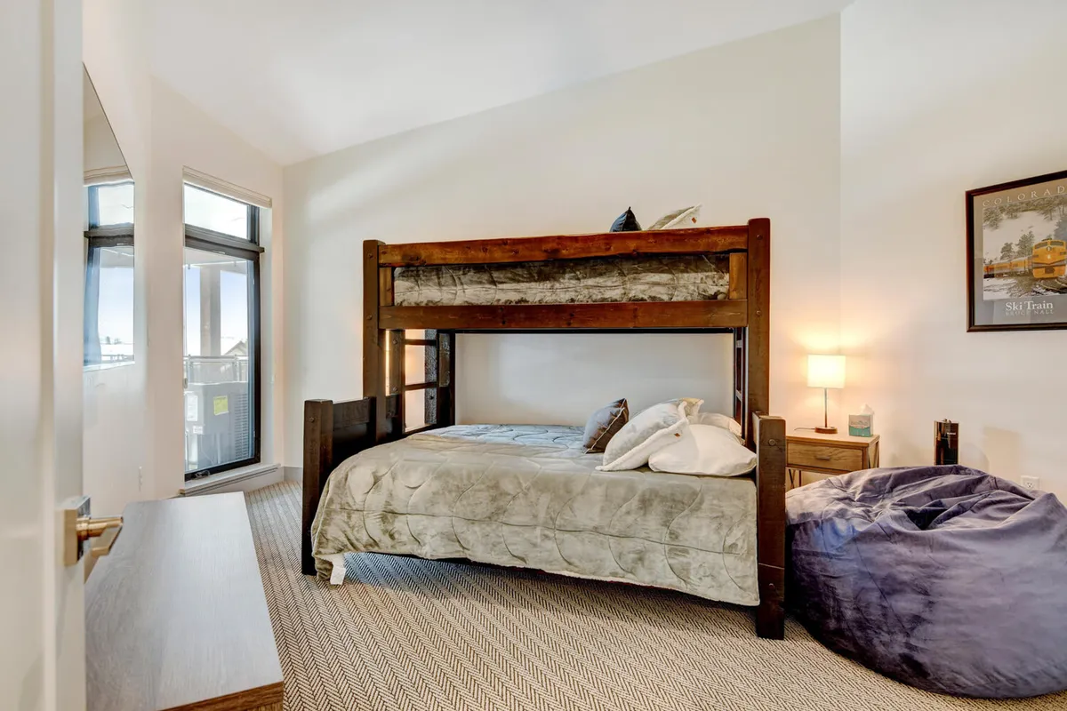 A playful, well-lit bedroom with a twin-over-queen bunk bed, bean bag chair, nightstand with lamp, and a window view. The room is designed for comfort and fun, making it an ideal space for children to sleep, read, and play.