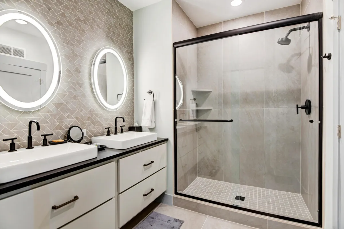 A modern, spa-inspired bathroom with double sinks, contemporary fixtures, a large walk-in glass shower, and elegant lighting. The space looks fresh, clean, and ideal for unwinding with self-care routines after a day out.