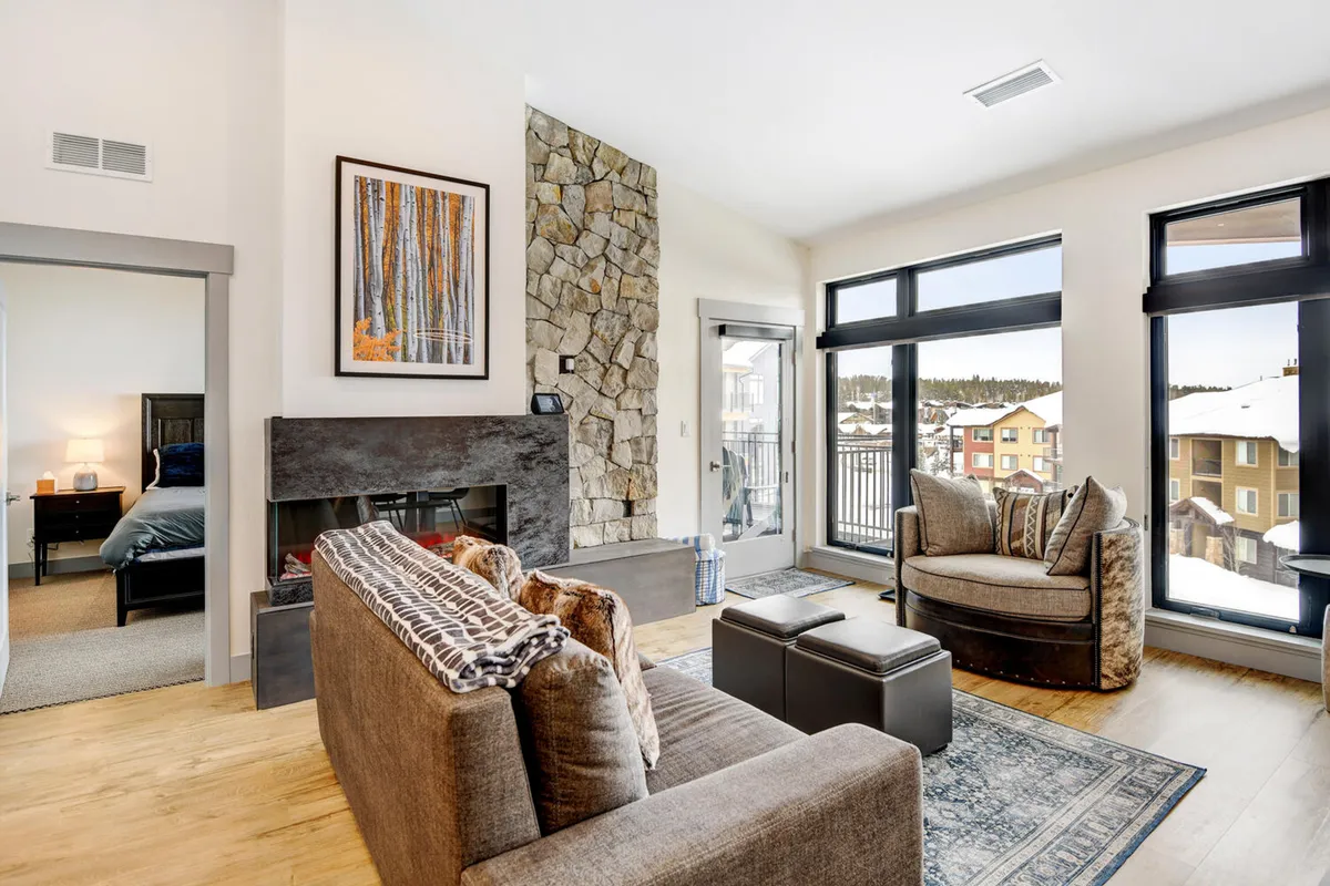 This image features a comfortable and welcoming living area with a stone fireplace, large windows that provide abundant natural light, stylish seating, and artwork on the wall. The windows frame a view of the outdoors, hinting at seasonal changes beyond the glass, while the cozy atmosphere inside makes it ideal for relaxing after a day of winter or summer adventures.
