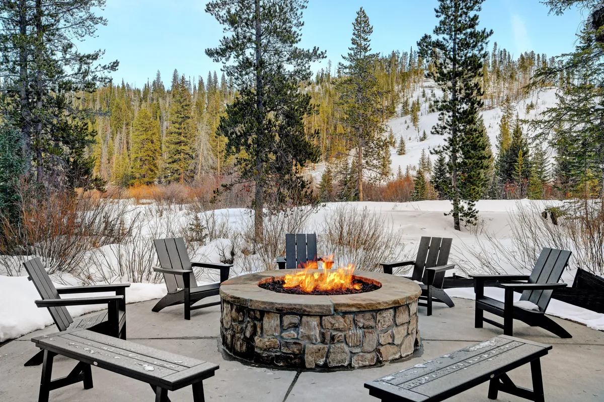An inviting fire pit is at the center of this snowy outdoor scene, surrounded by comfortable seating and tall trees. The snow-covered landscape and warm glow from the fire evoke a cozy, communal atmosphere, perfect for evening gatherings or storytelling after a day of adventure.
