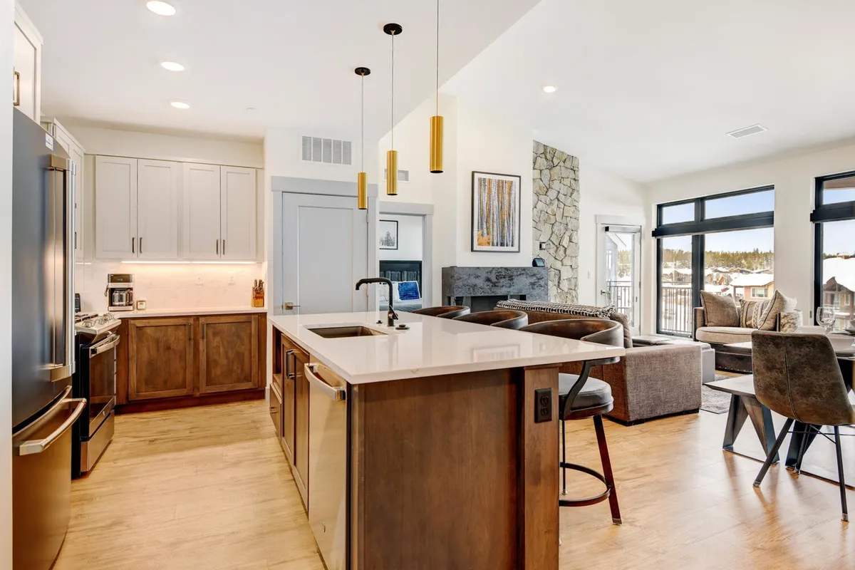 A bright, open-plan space featuring a kitchen island, stone fireplace, and large windows that look out onto the surrounding landscape. The area is designed for cooking, entertaining, and relaxing, with views that capture the ever-changing scenery outside, making it the perfect spot to gather before or after your favorite seasonal activities.