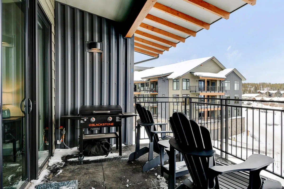 An exterior view of a patio area with a Blackstone grill and Adirondack chairs, framed by a balcony railing. The scene invites relaxation and outdoor gatherings, perfect for summer barbecues or winter cookouts.