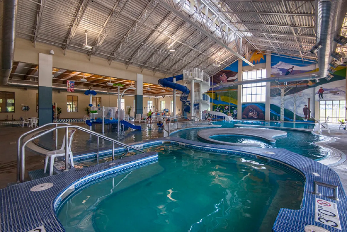 Bright indoor swimming pool area featuring a vibrant water slide, playful mural, and children enjoying the water. The scene bursts with energy and fun, highlighting family-friendly recreation and the excitement of the nearby Grand Park Rec Center.