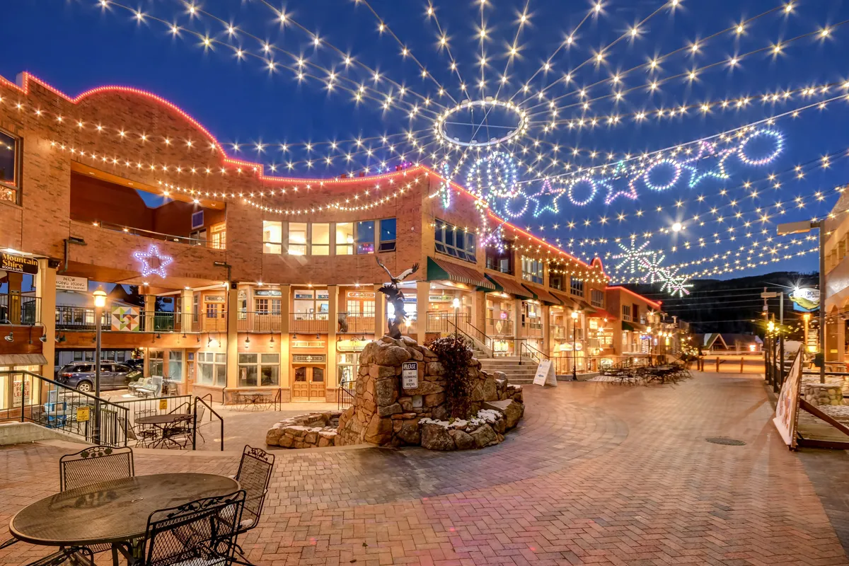 A charming outdoor plaza illuminated by string lights and adorned with decorative stars, featuring a stone fountain, inviting seating areas, and boutique shops. The scene exudes a joyful, celebratory mood, suggesting a space for holiday festivities and social gatherings.