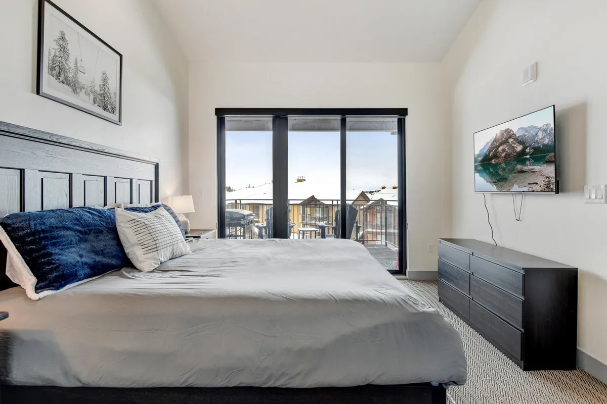 A spacious master suite featuring a large king-sized bed with crisp, high-quality linens, a mounted 50" HD TV on the wall, and expansive windows that frame stunning mountain views. The atmosphere is calm and serene, perfect for rest and relaxation after a day of adventure.