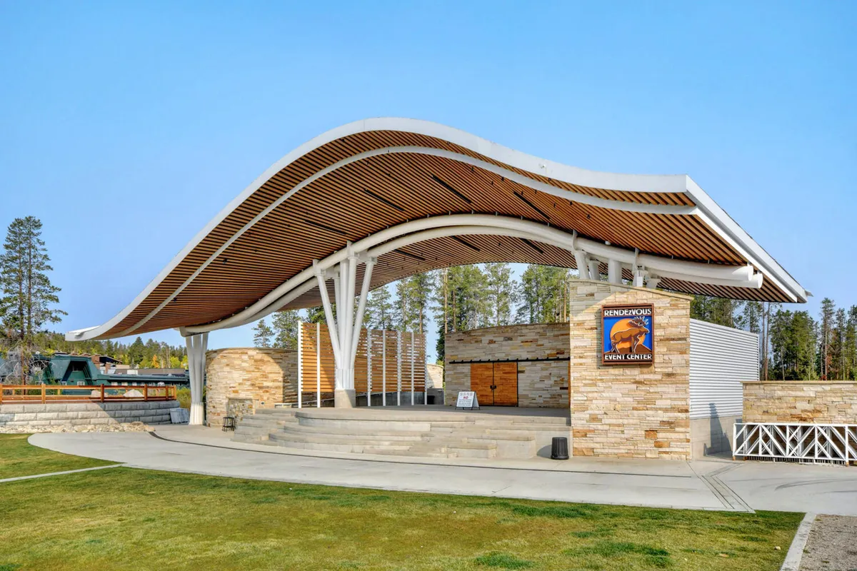 The Hideaway Park amphitheater stands as a modern outdoor venue with a distinct curved roof structure, stone walls, and a welcoming stage area nestled among lush greenery. The space is clearly designed for community gatherings, events, and performances in all seasons.