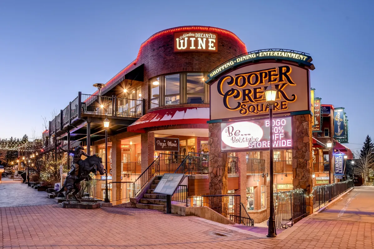 A lively downtown scene featuring the Cooper Creek Square sign, a wine shop, inviting outdoor seating areas, and a charming statue out front. The area is bustling and festive, with hints of live music and gatherings, capturing the atmosphere of Winter Park's social hub.