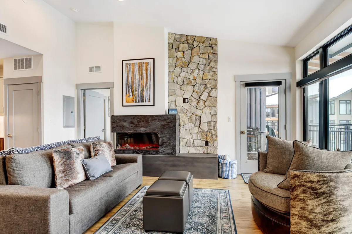 A sunlit living room featuring a striking stone fireplace, plush, inviting seating arranged for conversation or relaxation, and tasteful artwork that adds an upscale touch. The space exudes warmth and comfort, perfect for unwinding after mountain adventures or gathering with loved ones.