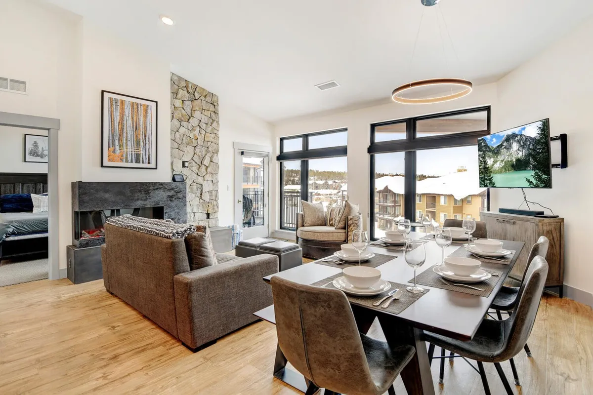 A spacious and open living area featuring a dining table set for a meal, a comfortable seating area with a couch, large windows offering scenic views, and a warm stone fireplace. The room is bathed in natural light, creating a welcoming atmosphere for gatherings and relaxation.