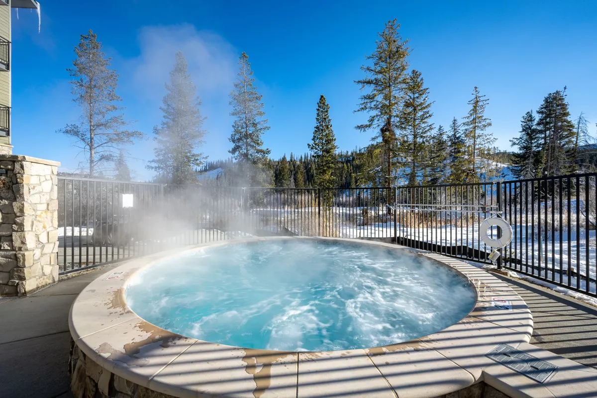 This exterior image captures the community hot tub surrounded by trees and an inviting fenced area. The tranquil scene is enhanced by natural elements, offering a sense of relaxation and privacy. People can be imagined relaxing in the steaming water, either at sunset or under the stars.